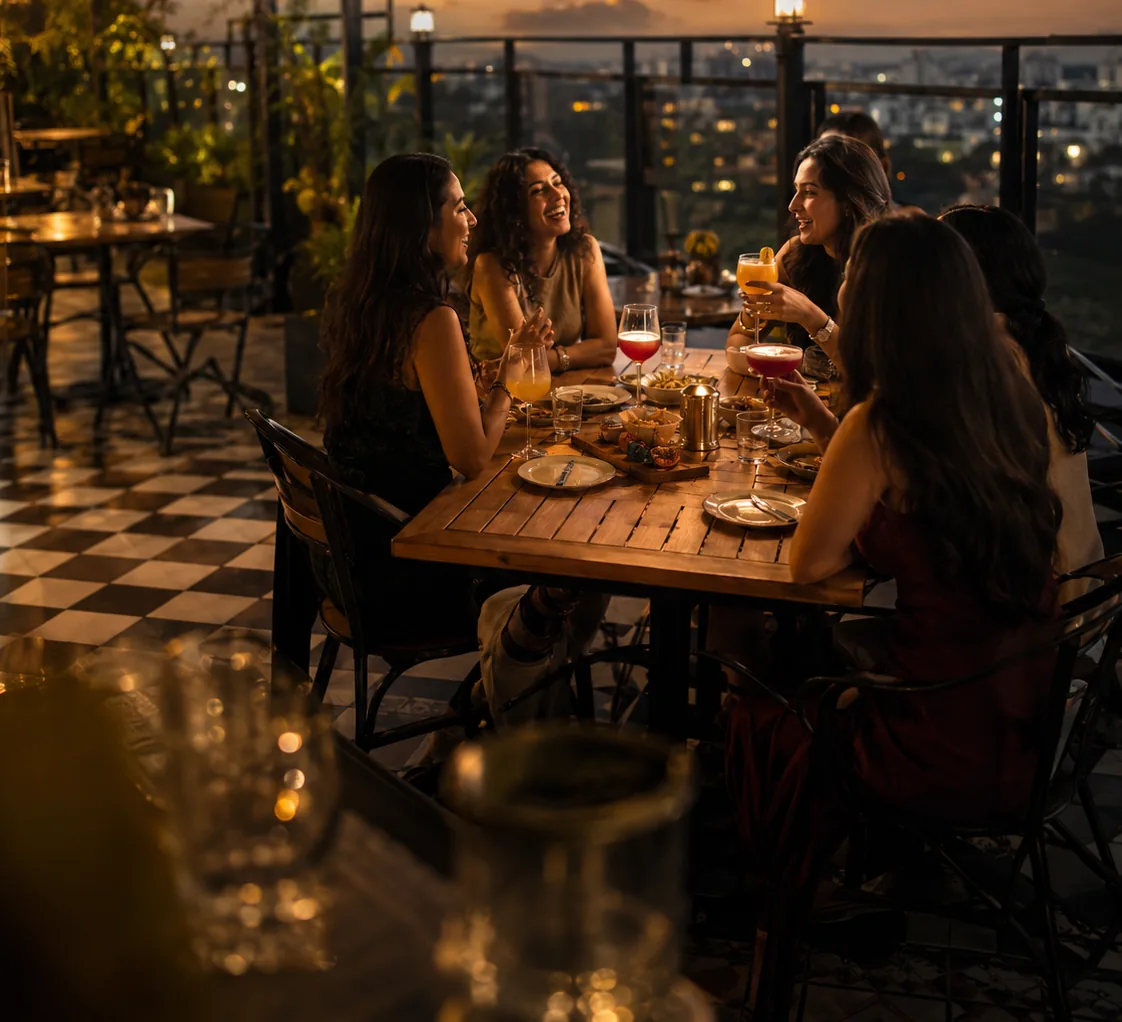 A group of women laughing and toasting cocktails together at a rooftop table at dusk, city skyline in the background